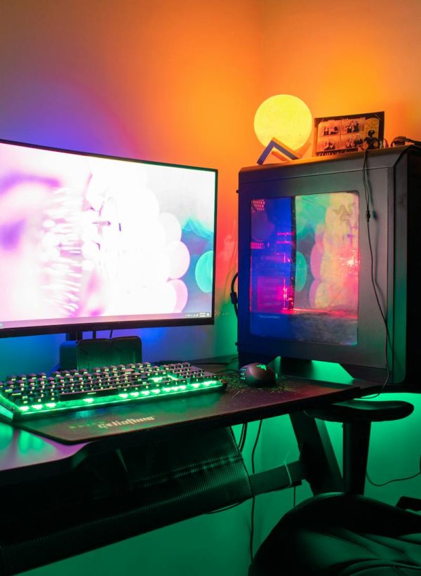 A colorful gaming setup featuring a PC, monitor, and gaming chair with RGB lights in a dark room.